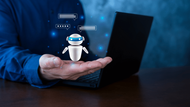 Hand houdt een kleine witte robot met chatbubbels vast boven een open laptop, tegen een blauwe achtergrond met lichtpuntjes
